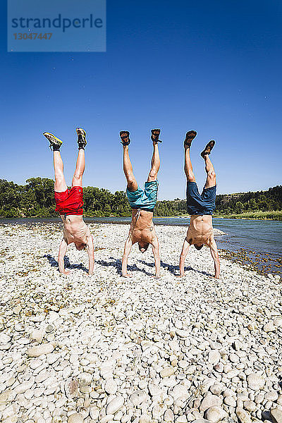 Freunde ohne Shirt machen Handstand am Seeufer vor klarem Himmel