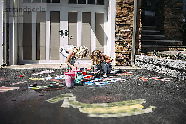 Verspielte Schwestern malen bei Sonnenschein auf Asphalt vor dem Haus