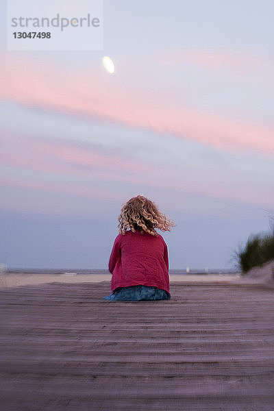 Rückansicht eines Mädchens  das bei Sonnenuntergang am Strand gegen den Himmel sitzt