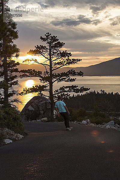 Mann fährt Skateboard auf Fußweg am See gegen den Himmel bei Sonnenuntergang