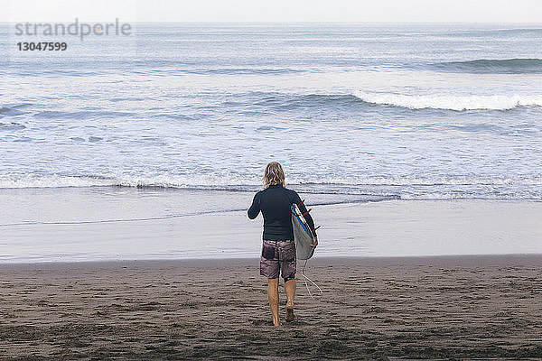 Hochwinkelansicht eines Mannes  der ein Surfbrett trägt  während er am Strand in Richtung Meer läuft