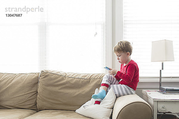 Seitenansicht eines Jungen  der ein Mobiltelefon benutzt  während er zu Hause auf dem Sofa sitzt