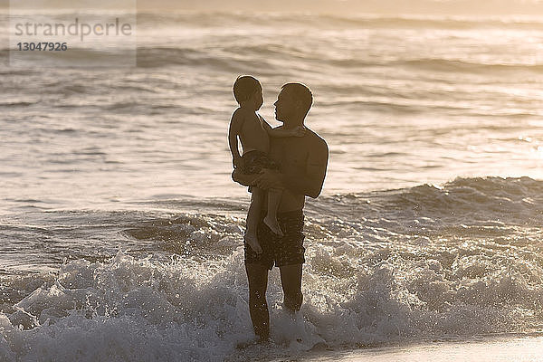 Vater ohne Shirt trägt Sohn beim Strandspaziergang bei Sonnenuntergang