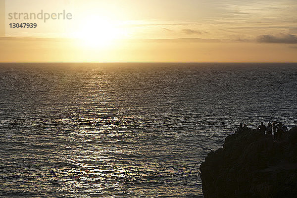 Silhouetten von Menschen  die bei Sonnenuntergang auf einer Klippe am Meer gegen den Himmel stehen