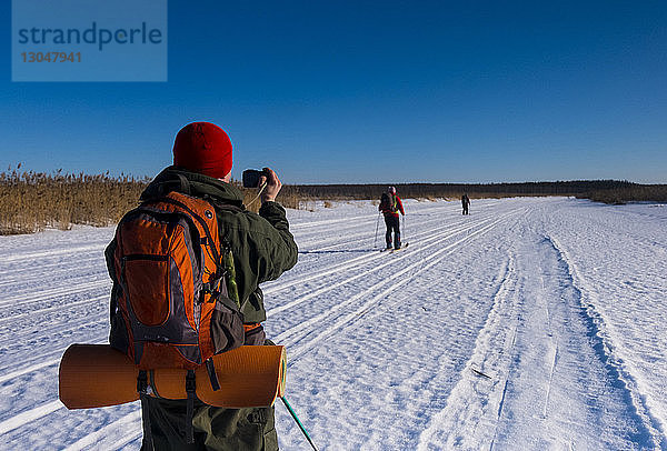 Rückansicht eines Mannes  der Freunde beim Skifahren auf schneebedecktem Feld vor strahlend blauem Himmel fotografiert