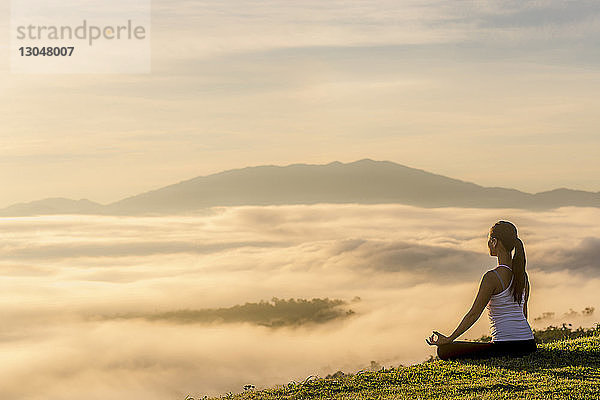 Seitenansicht einer Frau  die meditiert  während sie in einer Wolkenlandschaft auf dem Feld sitzt