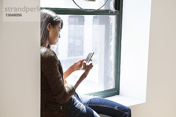 Seitenansicht einer Geschäftsfrau  die ein Smartphone benutzt  während sie im Büro auf dem Fensterbrett sitzt