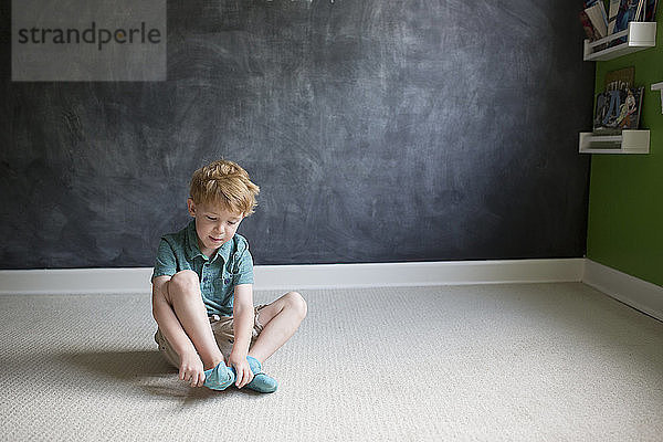 Junge in voller Länge trägt Socken  während er zu Hause auf Teppich gegen eine Tafelwand sitzt