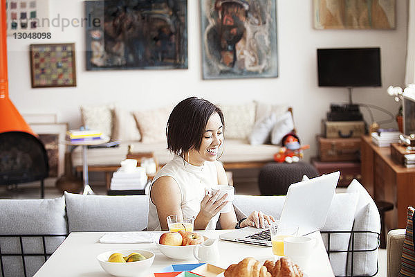 Glückliche Frau hält Kaffeetasse  während sie einen Laptop benutzt