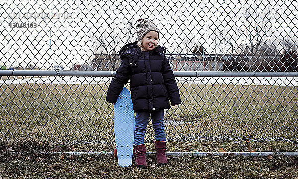 Lächelndes Mädchen mit Skateboard in der Hand  während es auf einem Grasfeld am Zaun des Parks steht