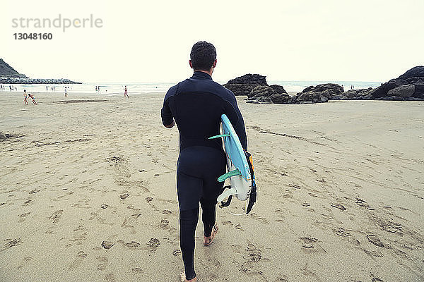 Rückansicht eines Mannes im Neoprenanzug mit Surfbrett beim Strandspaziergang