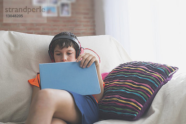 Junge benutzt Tablet-Computer  während er zu Hause auf dem Sofa Musik hört