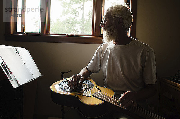 Älterer Mann schaut sich Papiere an  während er zu Hause Gitarre gegen Fenster lernt