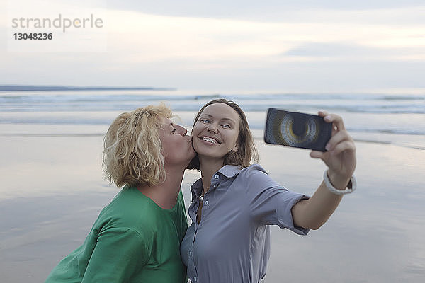 Fröhliche Frau  die sich mit einem Smartphone selbstständig macht  während sie bei Sonnenuntergang am Strand von einem Freund geküsst wird