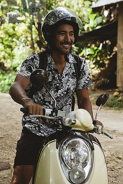 Lächelnder Mann mit Helm beim Fahren mit dem Motorroller auf unbefestigter Straße