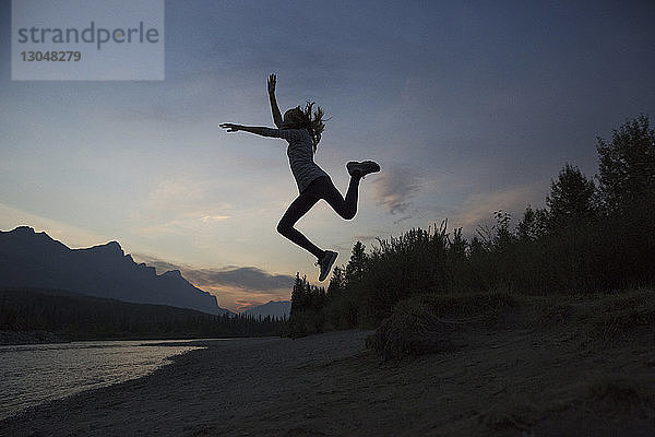 Silhouette eines glücklichen Teenagers  der im Banff-Nationalpark bei Sonnenuntergang am Seeufer gegen den Himmel springt