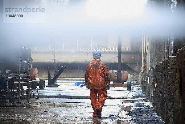 Rückansicht eines Handarbeiters mit Regenmantel und Helm beim Gang durch die Fabrik