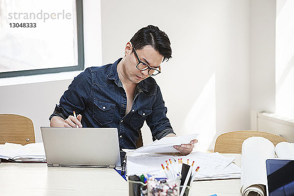 Geschäftsmann liest Dokument  während er mit seinem Laptop am Schreibtisch im Kreativbüro sitzt