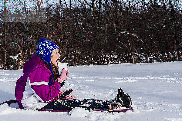 Seitenansicht eines Mädchens  das Schnee isst  während es auf einem Feld im Wald sitzt