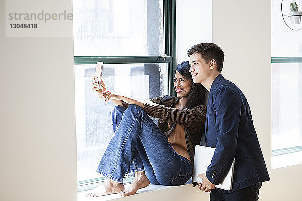 Fröhliche Geschäftskollegen  die sich im Kreativbüro am Fenster durch ein Smartphone selbstständig machen