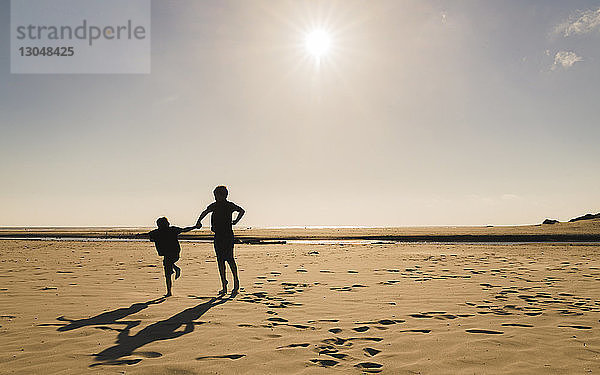 Scherenschnittbrüder springen bei Sonnenschein am Strand gegen den Himmel