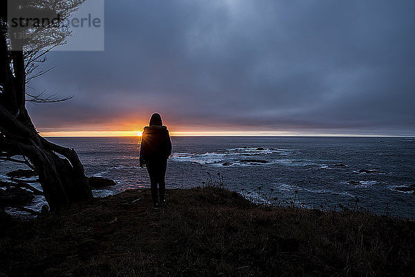 Silhouette eines Teenager-Mädchens  das bei Sonnenuntergang den Blick auf das Meer gegen den bewölkten Himmel richtet