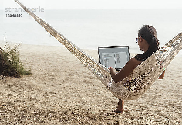 Frau benutzt Laptop-Computer  während sie sich auf Hängematte am Strand entspannt