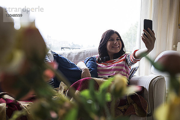 Lächelnde Frau  die sich mit dem Handy selbstständig macht  während sie sich an einem regnerischen Tag auf dem Sofa am Fenster entspannt