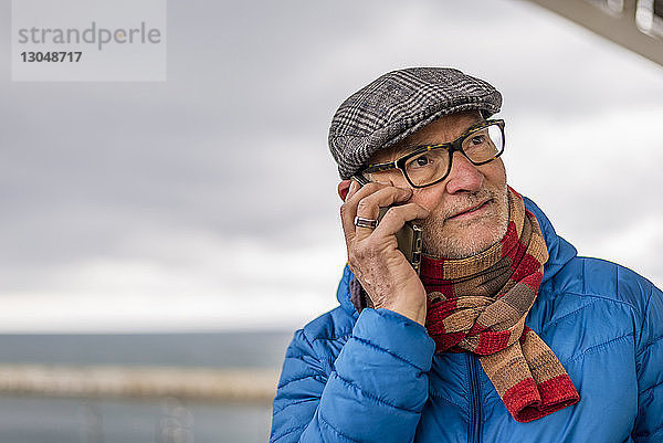 Nahaufnahme eines älteren Mannes  der mit einem Smartphone telefoniert  während er am Meer vor bewölktem Himmel steht