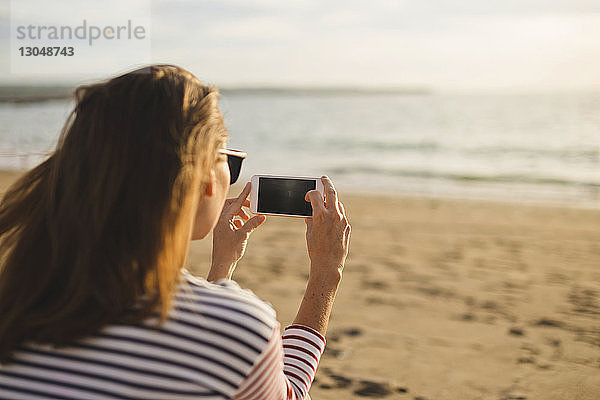 Rückansicht einer Frau  die das Meer fotografiert  während sie am Strand sitzt