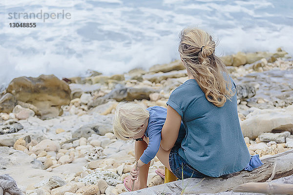 Rückansicht einer Mutter mit Sohn auf einem Baumstamm am Strand sitzend