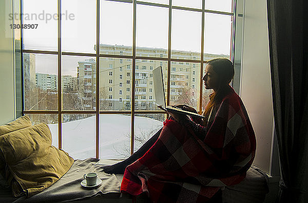 Seitenansicht einer Frau  die einen Laptop-Computer benutzt  während sie auf einem Fensterplatz in einer Nische sitzt