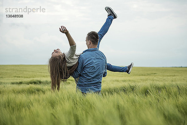 Rückansicht eines Mannes  der eine fröhliche Frau trägt  während er in einem Grasfeld gegen den Himmel steht