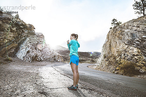 Seitenansicht einer Sportlerin in voller Länge  die die Arme ausstreckt  während sie auf einer Landstraße inmitten von Felsen steht