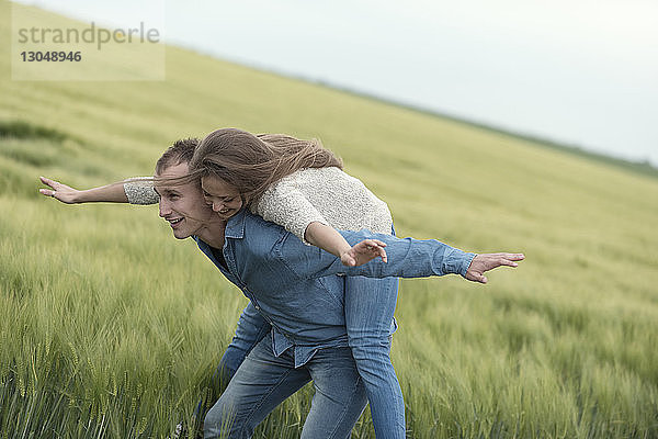 Fröhliches Paar mit ausgestreckten Armen genießt Huckepack-Ritt auf Grasfeld gegen den Himmel