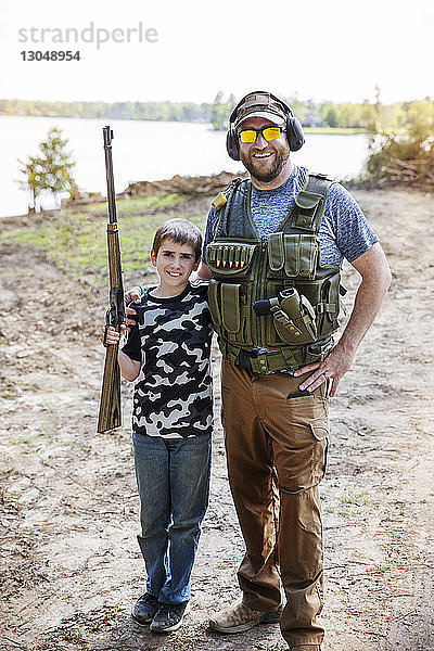 Porträt des Sohnes mit Gewehr in der Hand  während er mit dem Vater auf dem Feld gegen den Himmel steht