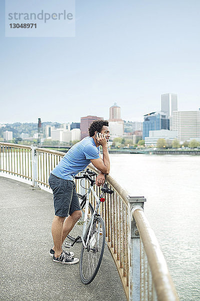 Männlicher Sportler  der ein Mobiltelefon benutzt  während er mit dem Fahrrad auf einer Brücke am Fluss steht