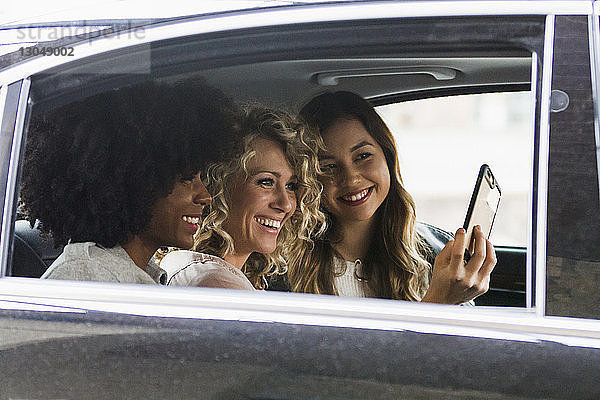 Fröhliche Geschäftsfrauen beim Selbermachen mit Smartphone im Auto durch Fenster gesehen