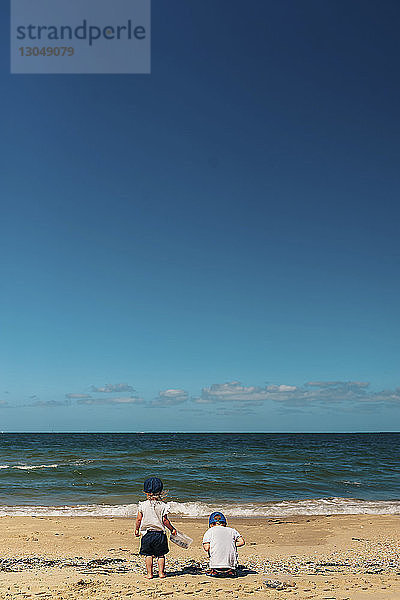 Rückansicht von Geschwistern beim Spielen am Strand vor blauem Himmel bei Sonnenschein