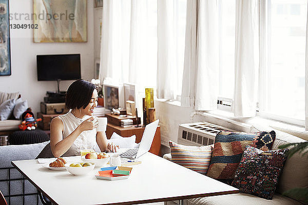 Glückliche Frau hält Kaffeetasse  während sie zu Hause einen Laptop benutzt