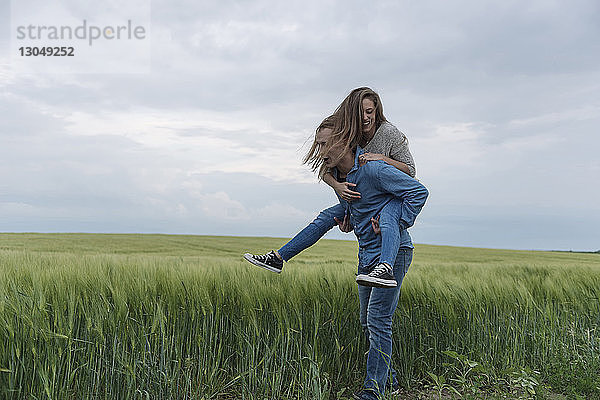 Junger Mann nimmt Frau huckepack auf Grasfeld vor bewölktem Himmel