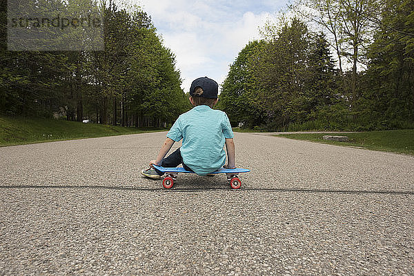 Rückansicht eines Jungen  der auf einem Skateboard am Fussweg sitzt