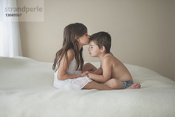 Mädchen küsst Bruder auf die Stirn  während sie zu Hause auf dem Bett sitzt