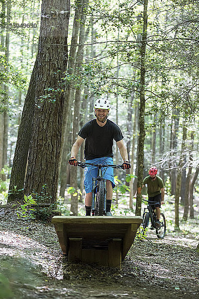 Glücklicher Wanderer macht Stunt beim Mountainbike fahren mit Freund im Hintergrund im Wald