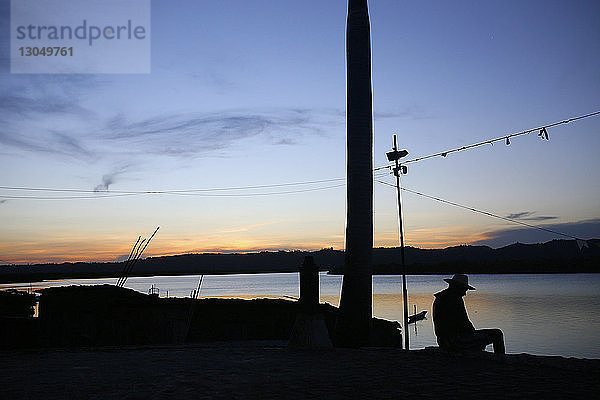 Scherenschnitt eines Mannes  der bei Sonnenuntergang am Seeufer gegen den Himmel sitzt