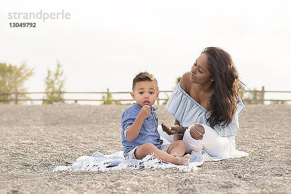 Porträt eines süßen Sohnes  der mit der Mutter auf einer Picknickdecke vor dem Himmel am Strand sitzt