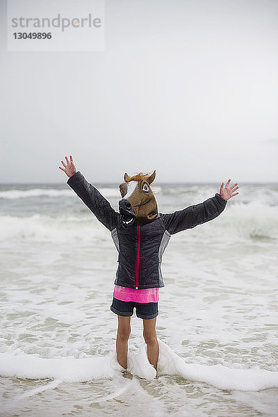 Verspieltes Mädchen mit Pferdemaske spielt am Strand vor klarem Himmel