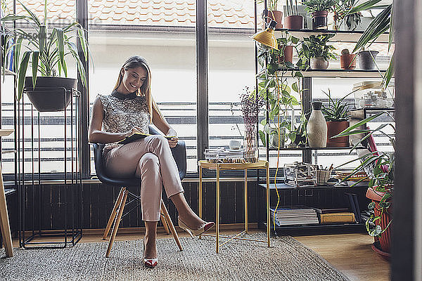 Glückliche Geschäftsfrau schreibt Tagebuch  während sie im Heimbüro auf einem Stuhl am Fenster sitzt