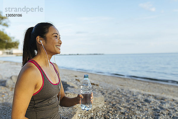 Glückliche Frau  die Musik hört  während sie eine Wasserflasche am Strand gegen den Himmel hält