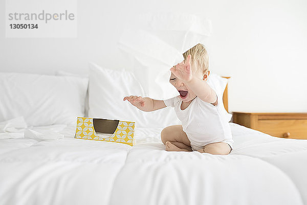 Fröhlicher Junge spielt zu Hause am Bett sitzend mit Gesichtstüchern